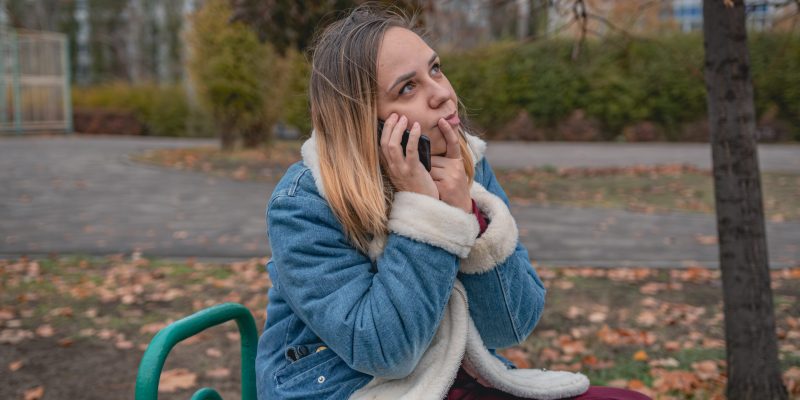 Young woman seated on a park bench conversing on her phone, surrounded by autumn leaves and trees in an urban setting